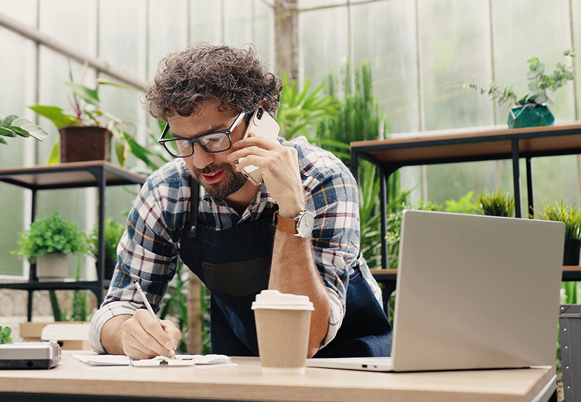 Business owner explores financial prospects over the phone with Landmark Member Services, considering a business checking account & credit card options. Member is taking notes at his store during the call.
