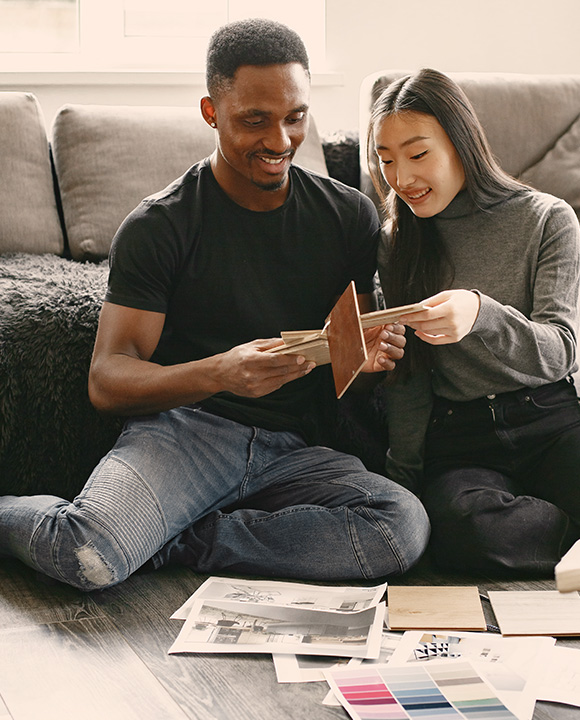A youthful couple sits on the floor of their residence, examining flooring samples, contemplating floor plan adjustments, and reviewing paint swatches.