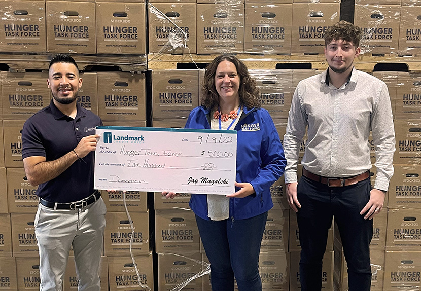 Landmark associates are proudly posing and holding a $500 check before a backdrop of Hunger Task Force boxes.