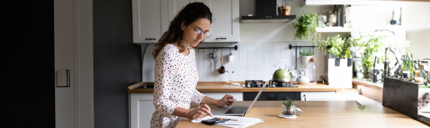Person in their kitchen is on their laptop with a calculator nearby looks where they can grow financially.
