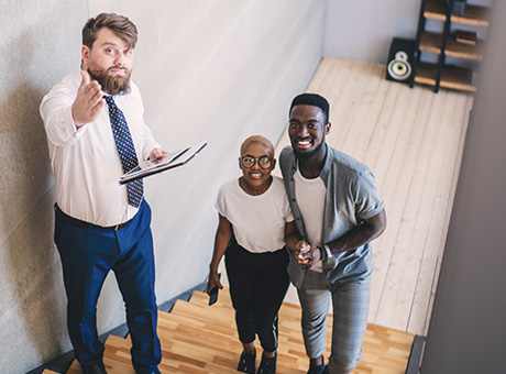 A realtor stands with an enthusiastic pair on a staircase, gesturing towards the second floor of their first home. 