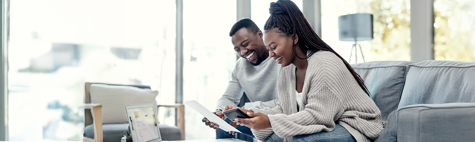 Couple sitting on a couch smiling looking at a smart phone and laptop
